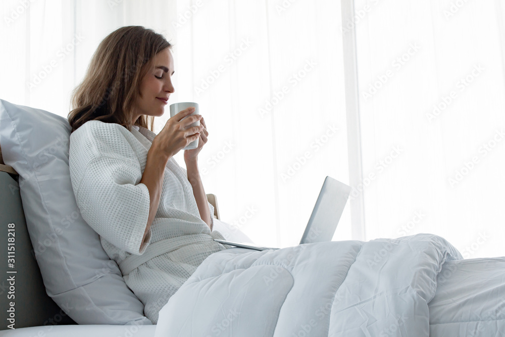Young caucasian woman in white pajamas with white holding a cup of coffee and drink and find information from the laptop on a bed. Young caucasian woman in white pajamas with white holding a cup of coffee and drink and find information from the laptop on a bed.