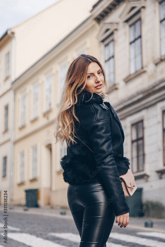 street style portrait of attractive blonde woman wearing leather trousers and jacket with a trendy purse. crossing the street and smiling at camera. fashion outfit perfect for autumn fall winter