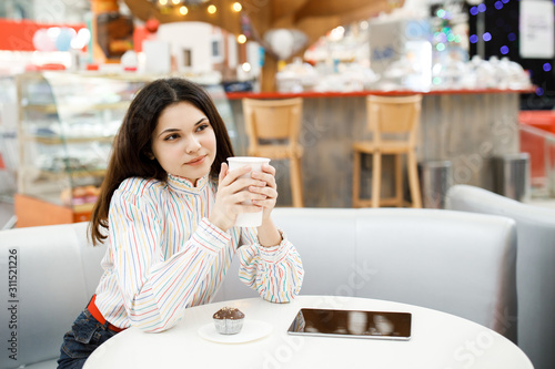 Pretty young brunette girl sitting in cafe and holding tablet, drinking coffee and resting