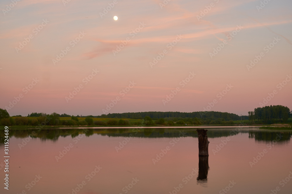 sunset on the shore of a pond with calm water, three cell towers and the moon in the sky.