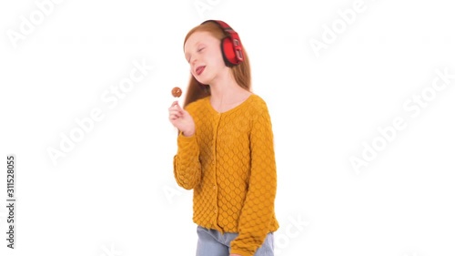 Close-up portrait shot of a lovely foxy haired girl wearing wireless headphones, eating lollipop candy while listening to the music. Isolated, on white background