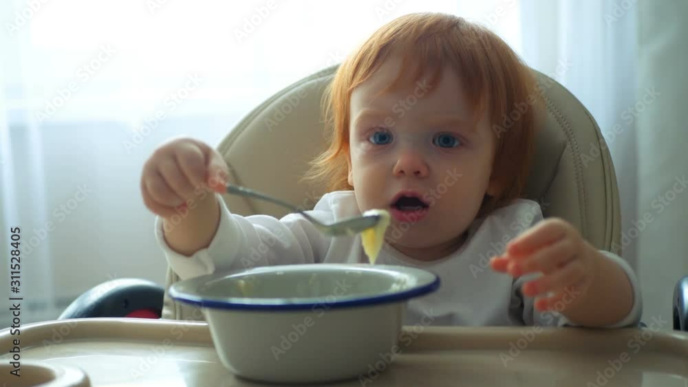 Close-up of a baby sitting at the table and eating baby food with a spoon on its own Close-up of a baby sitting at the table and eating baby food with a spoon on its own