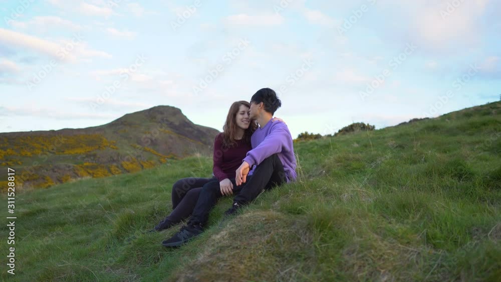 Young Couple Sitting on Grass in Outdoor Nature Scenery While Casually Chatting and Kissing Young Couple Sitting on Grass in Outdoor Nature Scenery While Casually Chatting and Kissing