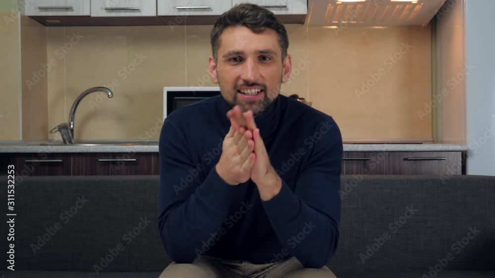 A young man in dark blue pullover and gray pants is sits on the couch, rubs his hands with pleasure and watches the TV show with interest. Kitchen on background. 4k slowmotion footage A young man in dark blue pullover and gray pants is sits on the couch, rubs his hands with pleasure and watches the TV show with interest. Kitchen on background. 4k slowmotion footage