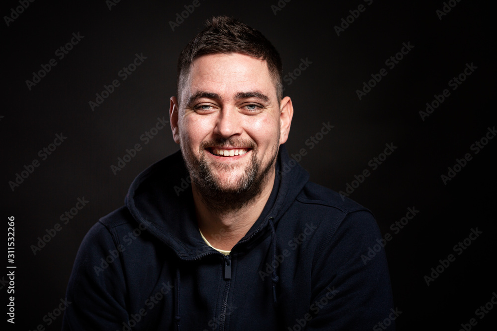 A young handsome smiling man in a blue hoodie is sitting and looking at the camera. Black background. Close-up.