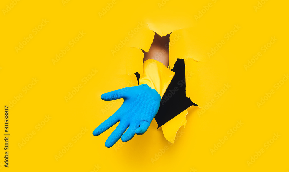 Fototapeta premium One male hand in yellow and blue household glove. Paper background and torn hole. Copy space. Guest worker concept.