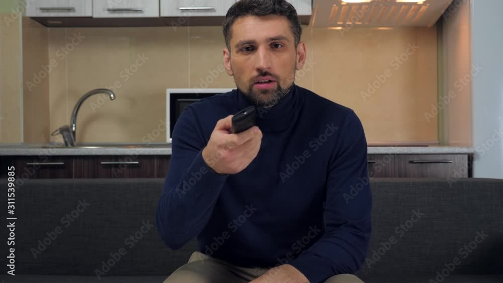 Young man in dark blue pullover and gray pants is sits down on couch, taking control remote in his hand, turns on the TV set and starts watching television show. Kitchen on background. 4k slowmotion Young man in dark blue pullover and gray pants is sits down on couch, taking control remote in his hand, turns on the TV set and starts watching television show. Kitchen on background. 4k slowmotion