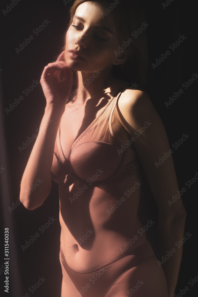 Naklejka premium sexy girl in lingerie touching face while posing in sunlight on black background