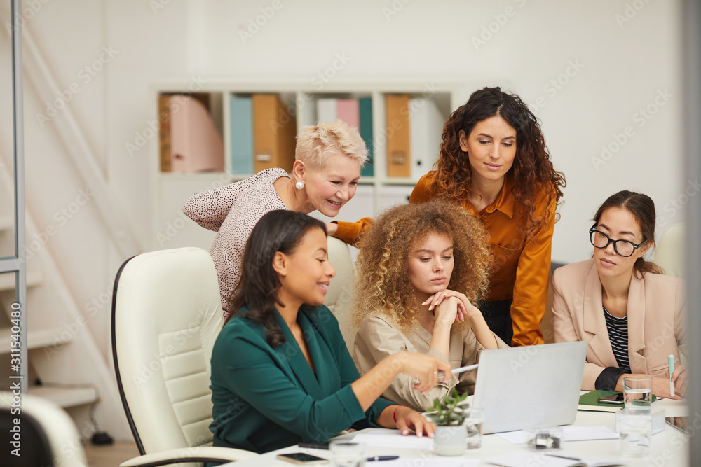 Fototapeta premium Group of five positive stylish women using laptop to work on project during business meeting horizontal shot