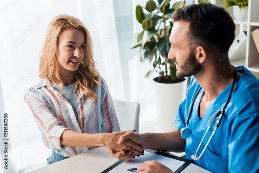 selective focus of happy woman shaking hands with handsome doctor in clinic selective focus of happy woman shaking hands with handsome doctor in clinic