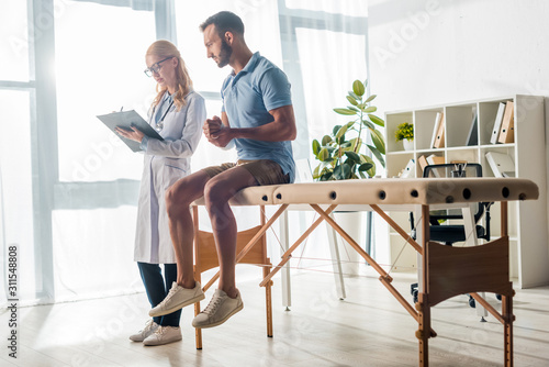 beautiful orthopedist writing diagnosis near injured bearded man