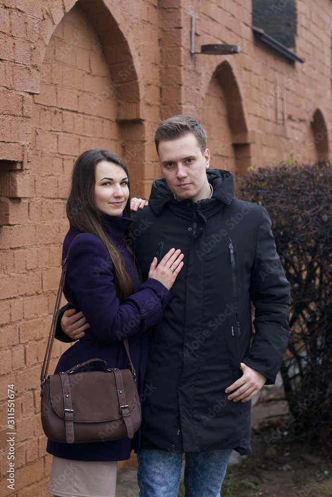 A young man hugs a girl at the waist. Against the background of a brown brick wall. Dressed in a coat and jacket.