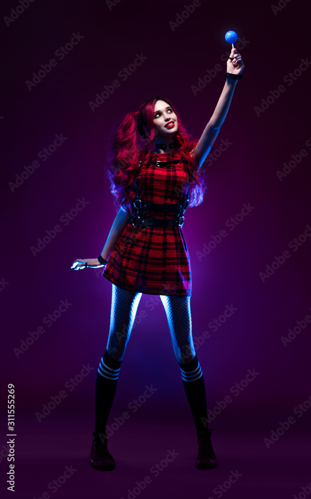 Attractive woman with red hair in checkered dress holding blue lollipop and posing on dark background