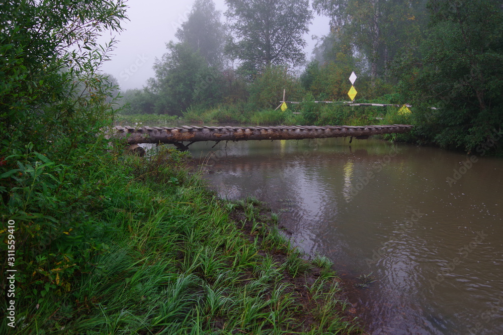  Old wooden bridge over the river. Morning. Fog. Reflection of a bridge in a river. Green bushes. Yellow grass. The dark color of water.