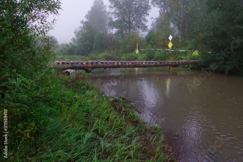  Old wooden bridge over the river. Morning. Fog. Reflection of a bridge in a river. Green bushes. Yellow grass. The dark color of water.