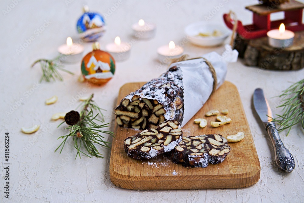 Dessert, chocolate salami with cookies and cashews in the New Year and Christmas style on a wooden board on a light concrete background. Desserts without baking. Dessert, chocolate salami with cookies and cashews in the New Year and Christmas style on a wooden board on a light concrete background. Desserts without baking.
