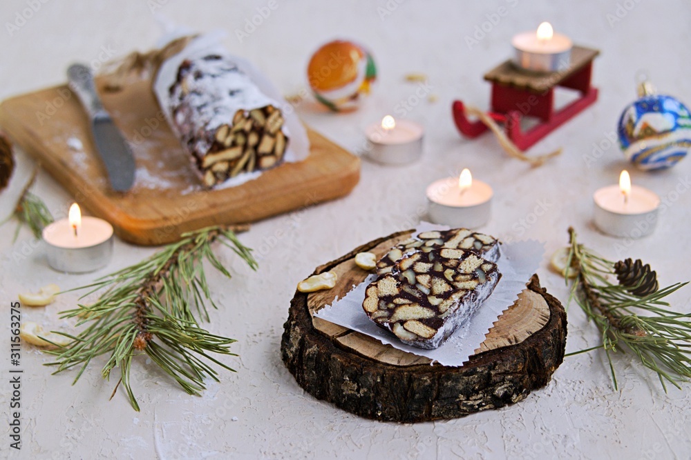 Dessert, chocolate salami with cookies and cashews in the New Year and Christmas style on a wooden board on a light concrete background. Desserts without baking. Dessert, chocolate salami with cookies and cashews in the New Year and Christmas style on a wooden board on a light concrete background. Desserts without baking.
