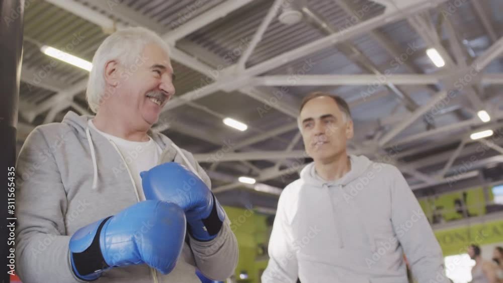 Close up of retired Caucasian sporty man wearing boxing gloves exercising with boxing bag while his middle-aged teammate standing nearby and watching. Then they making air punch Close up of retired Caucasian sporty man wearing boxing gloves exercising with boxing bag while his middle-aged teammate standing nearby and watching. Then they making air punch