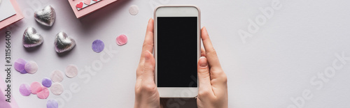 partial view of woman holding smartphone near valentines gifts and decoration on white background, panoramic shot