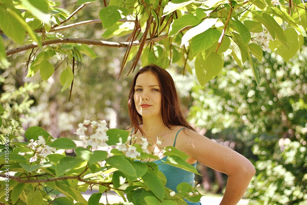 Beautiful brunette woman with long hair and blue eyes walking in the blooming park at spring in sunny day with bright sunshines and green trees Beautiful brunette woman with long hair and blue eyes walking in the blooming park at spring in sunny day with bright sunshines and green trees