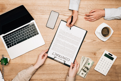 Top view of business partners sign contract by laptop, dollar banknotes and coffee on table, cropped view