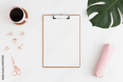 Top view of a wooden clipboard mockup with  workspace accessories, coffee and a monstera leaf on a white table.