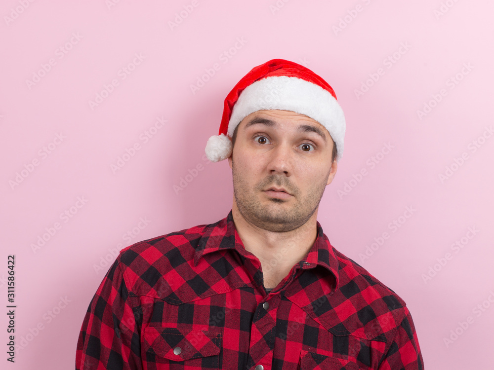 Emotions on the face, tired, holiday hangover, awareness. A man in a plaid rabbit and a Christmas red hat, on a pink background, copy space. Emotions on the face, tired, holiday hangover, awareness. A man in a plaid rabbit and a Christmas red hat, on a pink background, copy space.