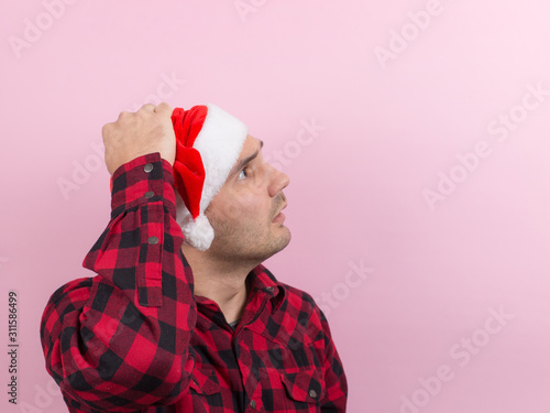 Emotions on the face, fear, holiday memories, negative. A man in a plaid rabbit and a Christmas red hat, on a pink background, copy space.