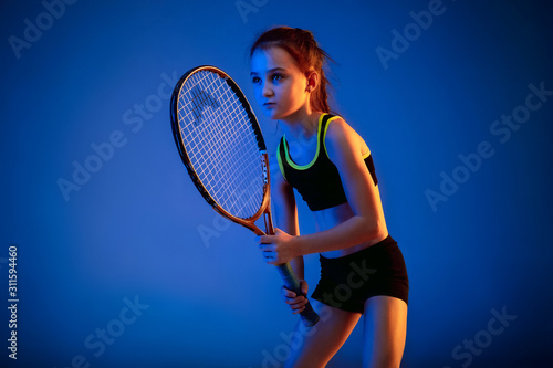 Little caucasian girl playing tennis on blue studio background in neon light. Cute model training in motion, action. Youth, flexibility, power, energy. Movement, ad, sport, healthy lifestyle concept. © master1305