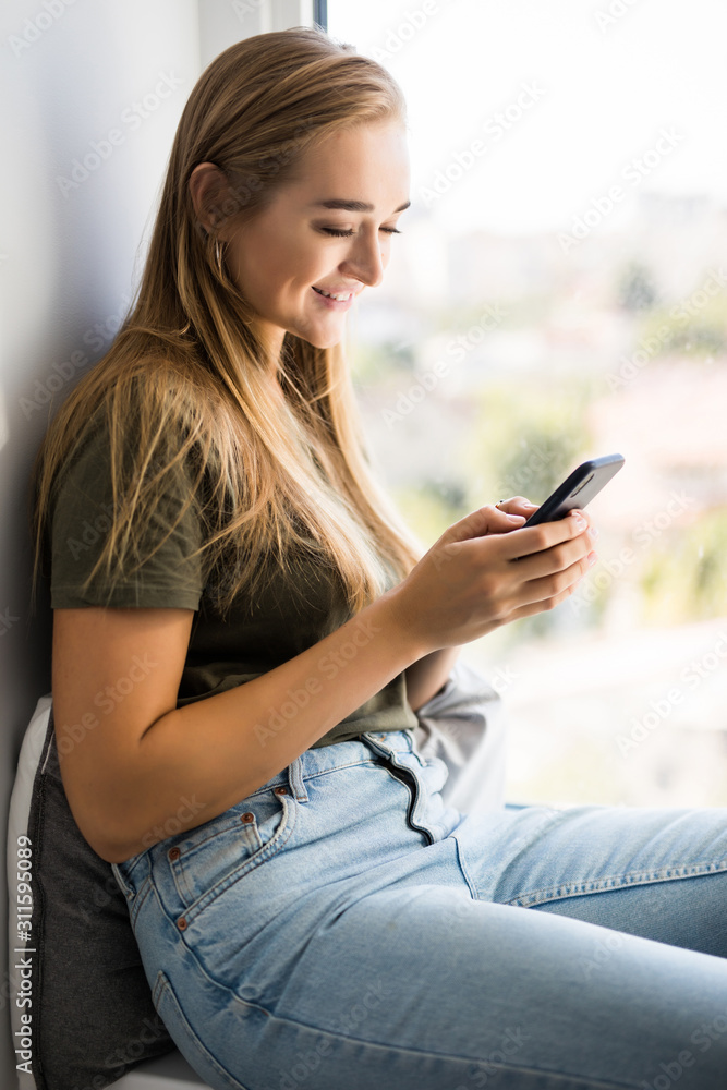 Gorgeous young woman checking email and sending sms message on mobile phone sitting on windowsill Gorgeous young woman checking email and sending sms message on mobile phone sitting on windowsill