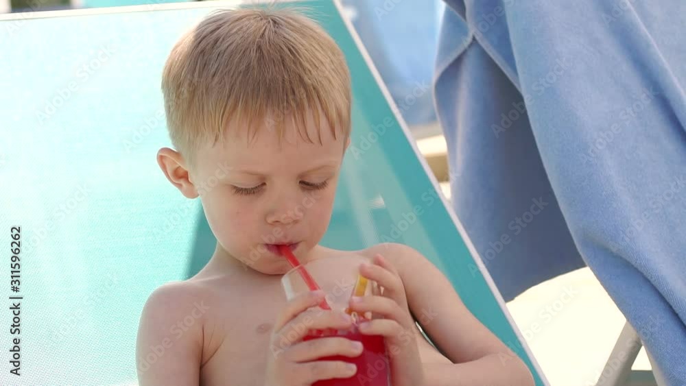 Close-up of a small blond boy drinking a cocktail by the pool in the summer in hot Sunny weather. Slow motion. Close-up of a small blond boy drinking a cocktail by the pool in the summer in hot Sunny weather. Slow motion.