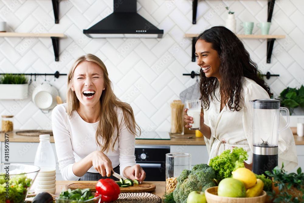 Two beautiful young girls are making a healthy breakfast and sincerely laughing near the table full of fresh vegetables on the white modern kitchen - obrazy, fototapety, plakaty 