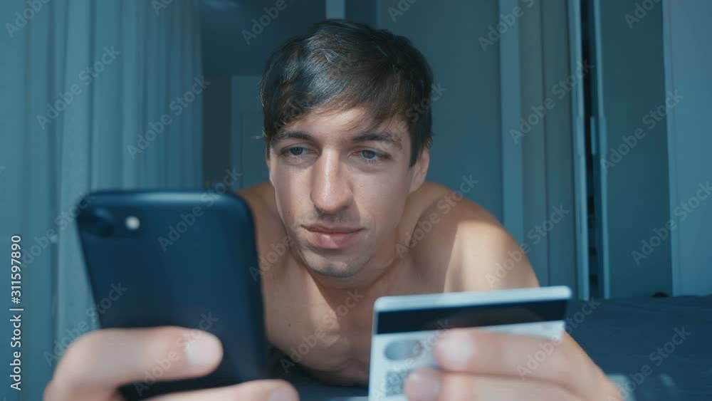 Naked man buying online, using smartphone and a credit card at home lying in bed, shopping cellphone app. Happy male holding credit card, buying online, using smartphone. Naked man buying online, using smartphone and a credit card at home lying in bed, shopping cellphone app. Happy male holding credit card, buying online, using smartphone.