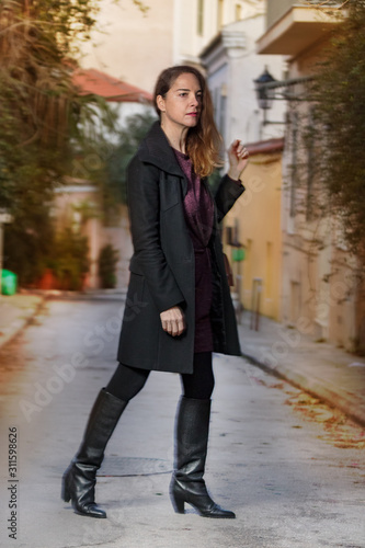 Full-length fashion portrait of a mature woman wearing high knee black boots, camel shoulde bag and a black slim fit coat, feeling cold, blue night.