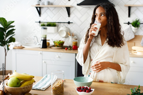 Smiled attractive girl is drinking milk near the table with fresh fruits on white modern kitchen dressed in nightwear with loose hair and looking on the right