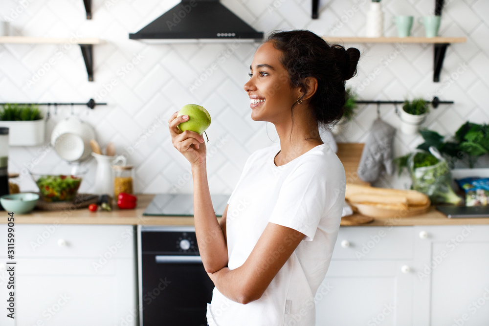 Lateral view of smiled attractive girl who is holding an apple and looking far away on the white modern kitchen Lateral view of smiled attractive girl who is holding an apple and looking far away on the white modern kitchen