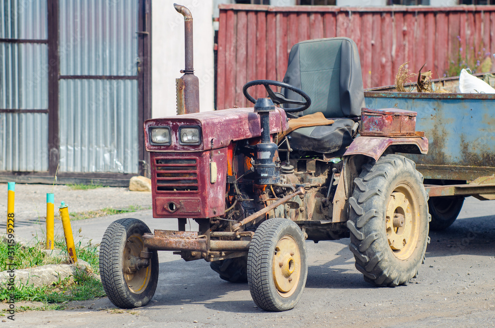 A small old mini tractor with a trailer stands on a rural street A small old mini tractor with a trailer stands on a rural street