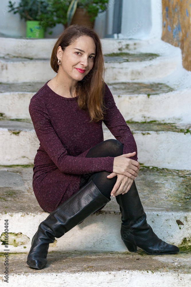 Full length fashion portrait of a mature woman wearing high knee black boots and a purple dress sitting outdoors. Adobe Stock
