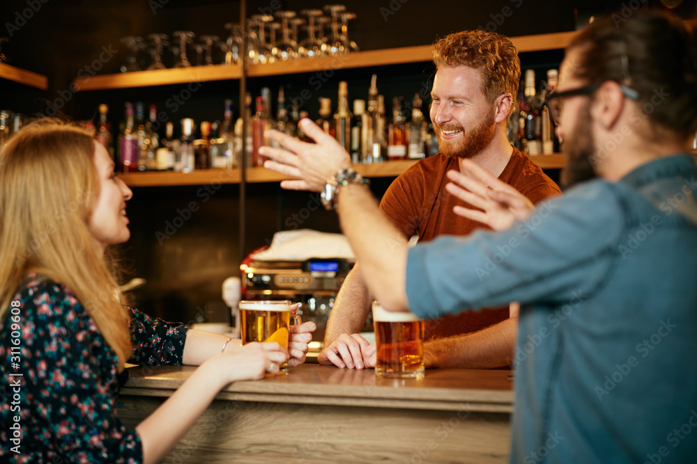 Cheerful friends leaning on bar counter, drinking beer and chatting with bartender. Night out.
