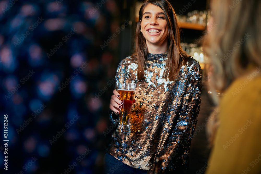 Beautiful caucasian smiling stylish woman holding beer and chatting with friends. In background is bar counter. Pub interior. Beautiful caucasian smiling stylish woman holding beer and chatting with friends. In background is bar counter. Pub interior.