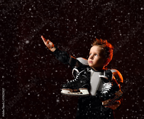 Serious boy in protective hockey uniform with skates on neck standing and reaching up over dark background with snowflakes