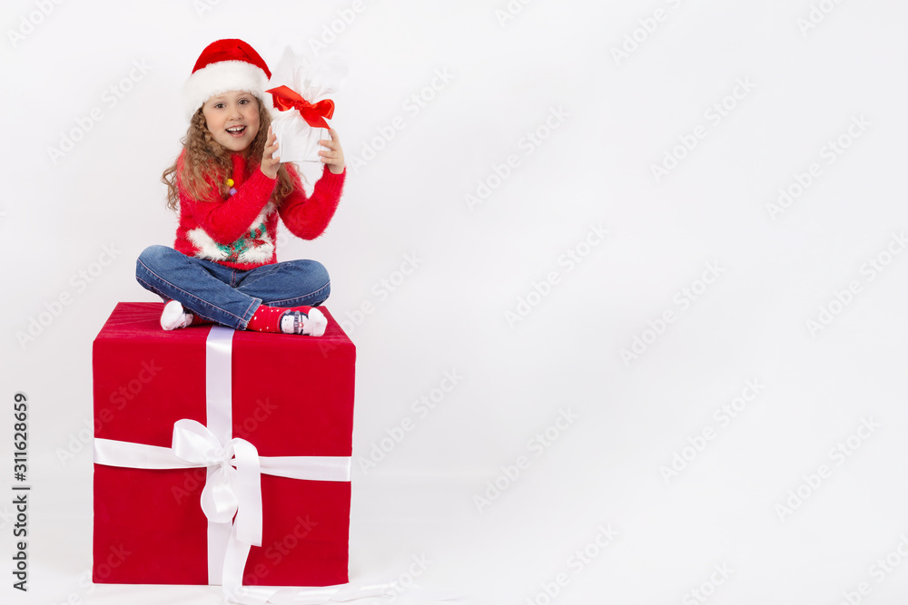 A little girl in a Santa Claus hat sitting on a red cube in the Studio with a gift in her hands, on a white background. The concept of a happy childhood. Blank space for text. The layout for the cards A little girl in a Santa Claus hat sitting on a red cube in the Studio with a gift in her hands, on a white background. The concept of a happy childhood. Blank space for text. The layout for the cards
