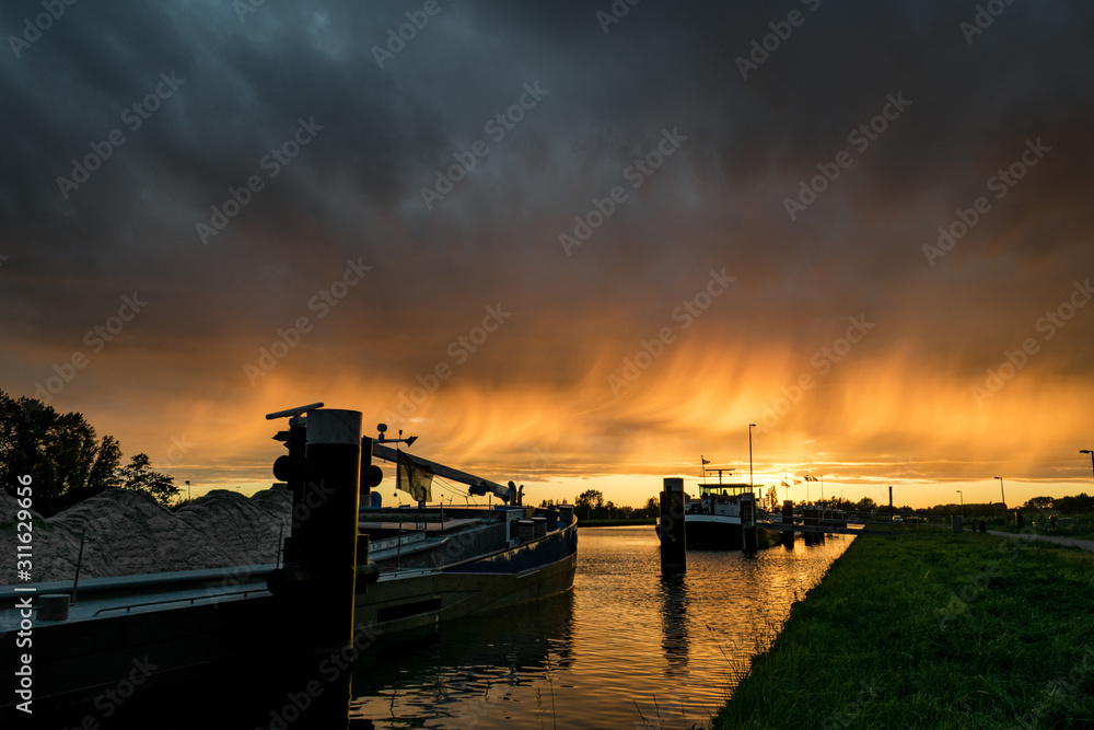 Very colorful sunset where the falling rain is illuminated by the setting sun. Boats in a small harbor in the foreground.