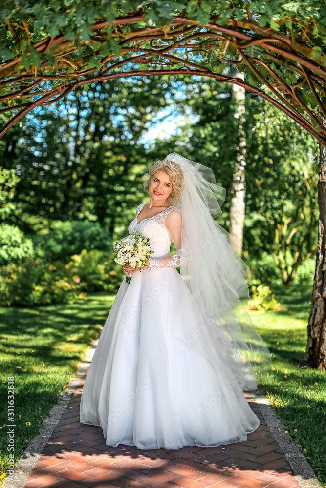 Beautiful blonde bride stands near the arch in greenery. Wedding portrait of a cute bride with a bouquet in the scenery. Concept and photography.