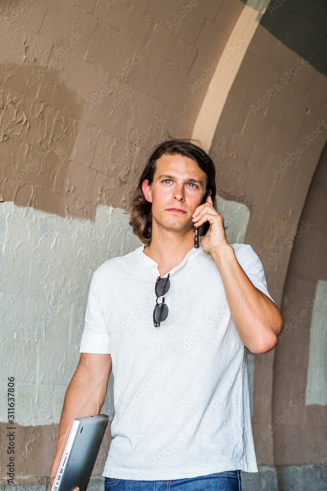 Young American Man with long hair traveling, working in New York City,  wearing white shirt, sunglasses hanging on collar, carrying laptop  computer, walking under street bridge, talking on cell phone.. Stock Photo