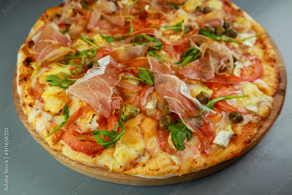 The pizza is prosciutto. Capers, tomatoes, arugula, mozzarella, brie cheese. Wooden plate. Gray background. Restaurant The pizza is prosciutto. Capers, tomatoes, arugula, mozzarella, brie cheese. Wooden plate. Gray background. Restaurant