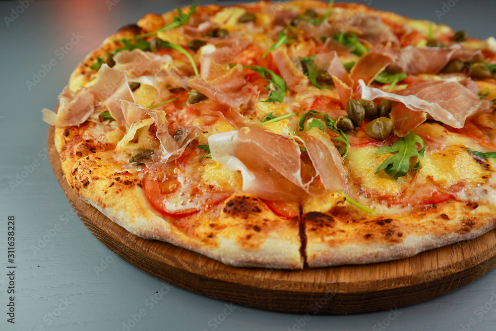 The pizza is prosciutto. Capers, tomatoes, arugula, mozzarella, brie cheese. Wooden plate. Gray background. Restaurant