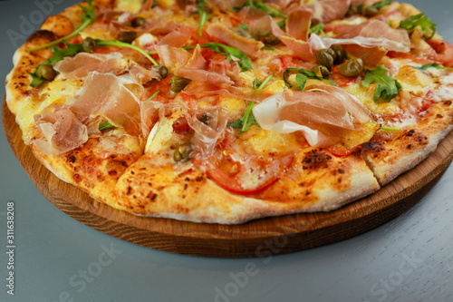 The pizza is prosciutto. Capers, tomatoes, arugula, mozzarella, brie cheese. Wooden plate. Gray background. Restaurant