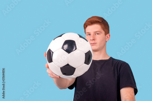 Shot of a red-haired teenager holding a soccer ball in his hand on a blue background.