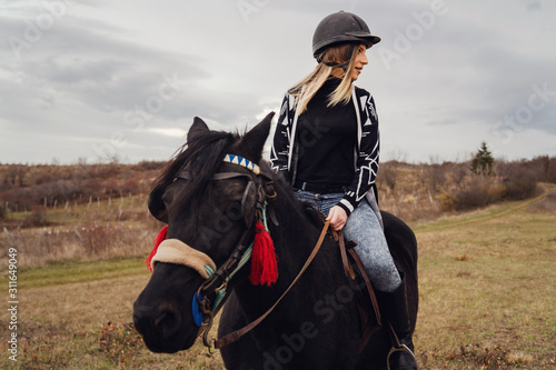 Young beautiful blonde caucasian woman riding a horse horseback casual rider girl in autumn or winter day wearing protective helmet © Miljan Živković